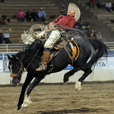 photos ©Bill Patterson, Patterson Photography a photo of a cowboy in a red shirt and high boots riding a bucking black horse in a rodeo arena